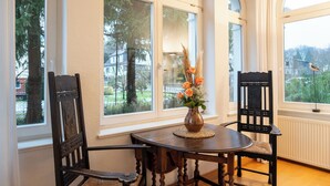 A room with a round table, two chairs, a vase with flowers, and large arched windows with a view of trees and buildings. - Villa Cremona (Seebad Bansin)