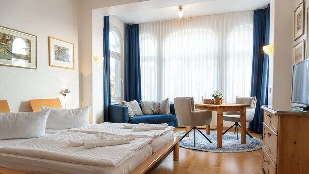 A hotel room with a large bed, a sofa, a dining table with chairs, a TV, and a dresser. - Villa Cremona (Seebad Bansin)