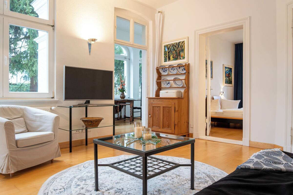 A living room with a glass coffee table, white sofa, and a TV. There is a wooden cabinet and a painting on the wall.