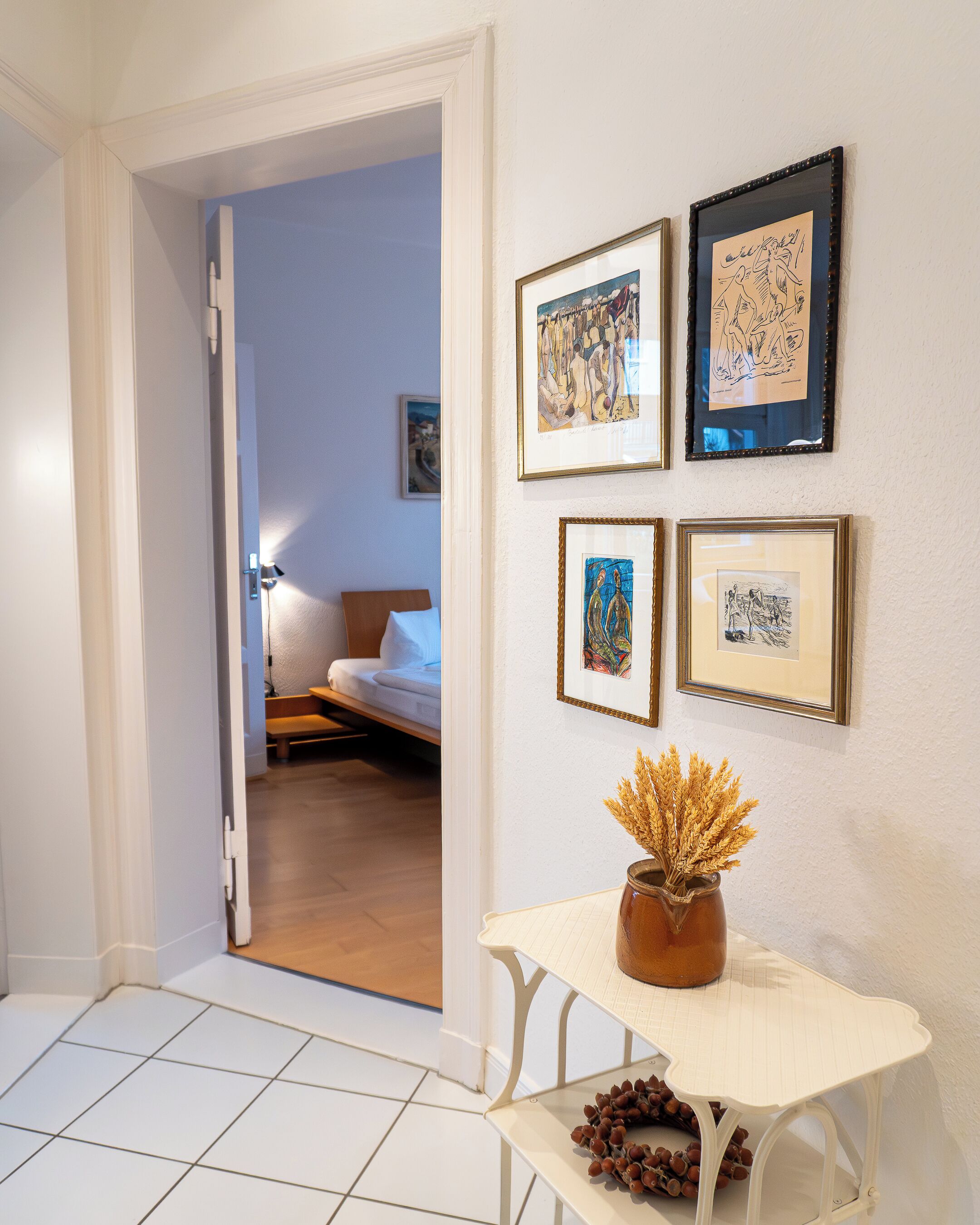 A hotel room with a bed, a small table with a vase and pine cones, and four framed artworks on the wall.
