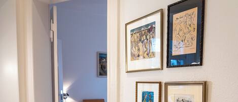 A hotel room with a bed, a small table with a vase and pine cones, and four framed artworks on the wall.