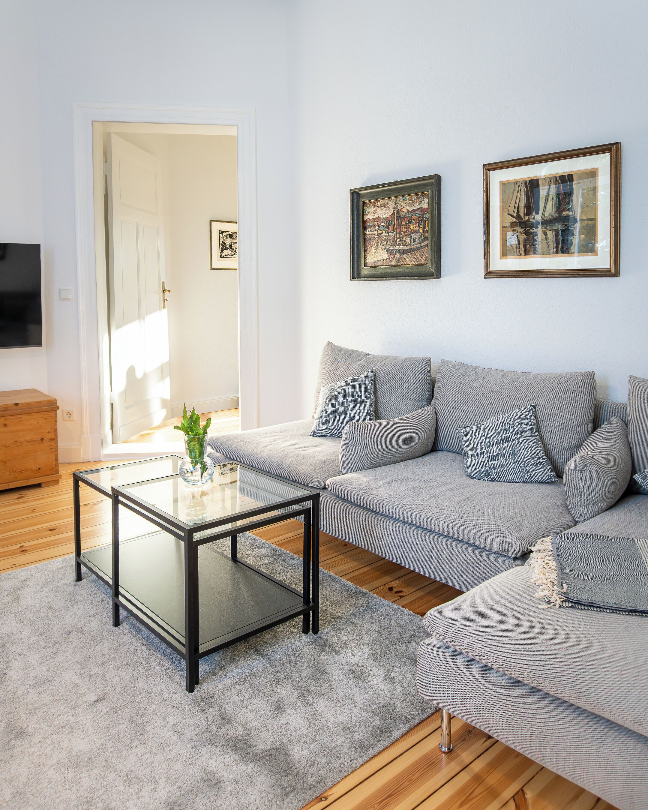 A modern living room with a grey sofa, a glass coffee table, and two framed artworks on the wall.