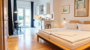 A modern hotel room with a wooden bed, a dining area with a table and chairs, a kitchen area, and a large window with curtains. - Villa Cremona (Seebad Bansin)