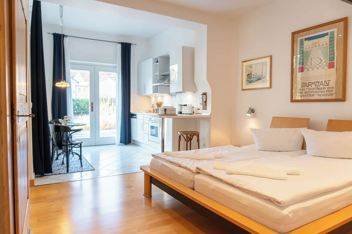 A modern hotel room with a wooden bed, a dining area with a table and chairs, a kitchen area, and a large window with curtains.