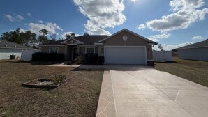 A single-story house with a garage, a front yard with a small garden, and a clear blue sky with clouds. - Spacious 4-bedroom house in perfect Ocala with cool AC (Ocala)