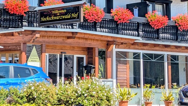 A multi-story building with balconies adorned with red flowers, a sign reading "Willkommen im Hochschwarzwald-Hof," and a German sign indicating a German restaurant.