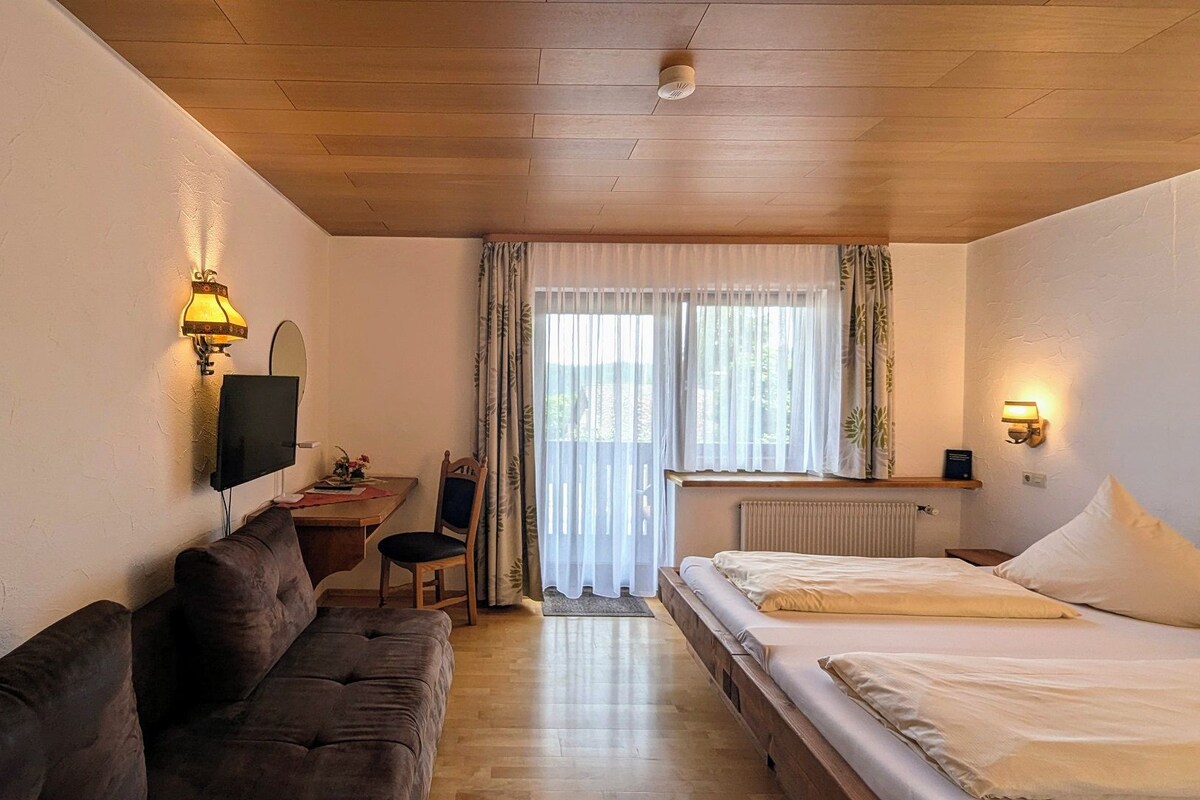 A hotel room with a wooden ceiling, a sofa, a bed, a desk, a chair, and a window with curtains.
