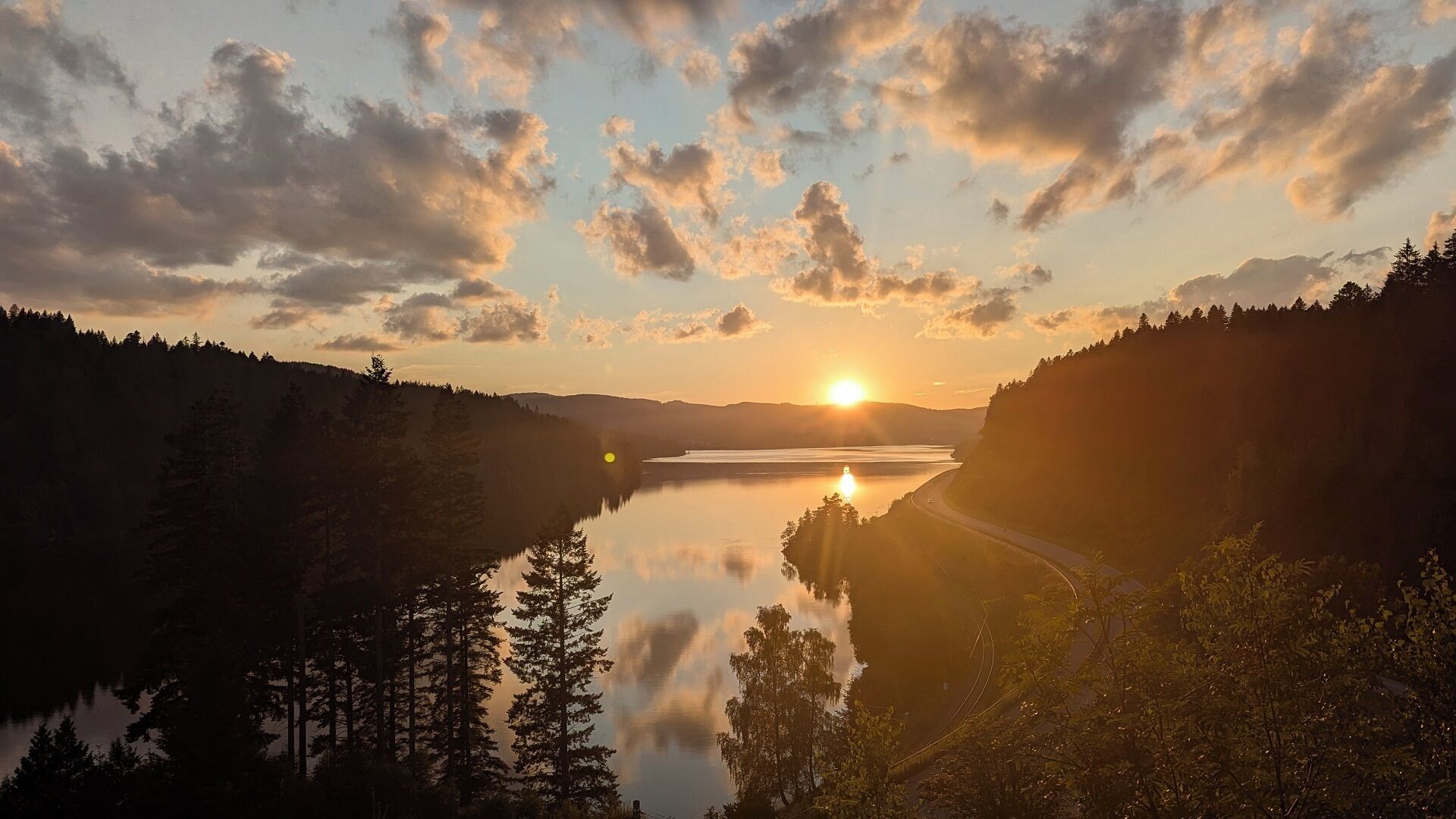 A serene landscape with a lake, forested hills, and a road leading towards the sunset.