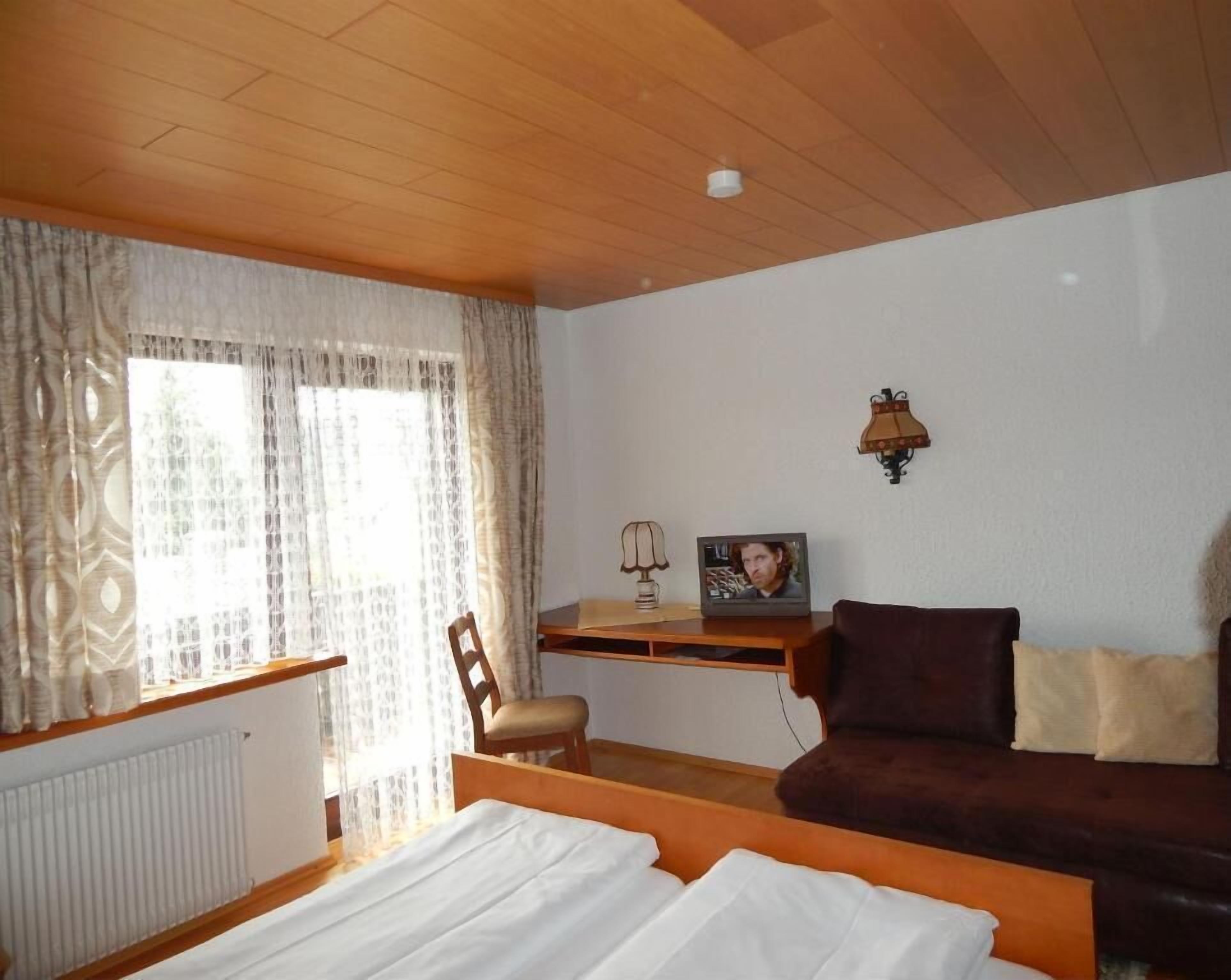 Habitación de hotel con techo de madera, cama con ropa blanca, sofá marrón, pequeña mesa con lámpara y un cuadro en la pared.