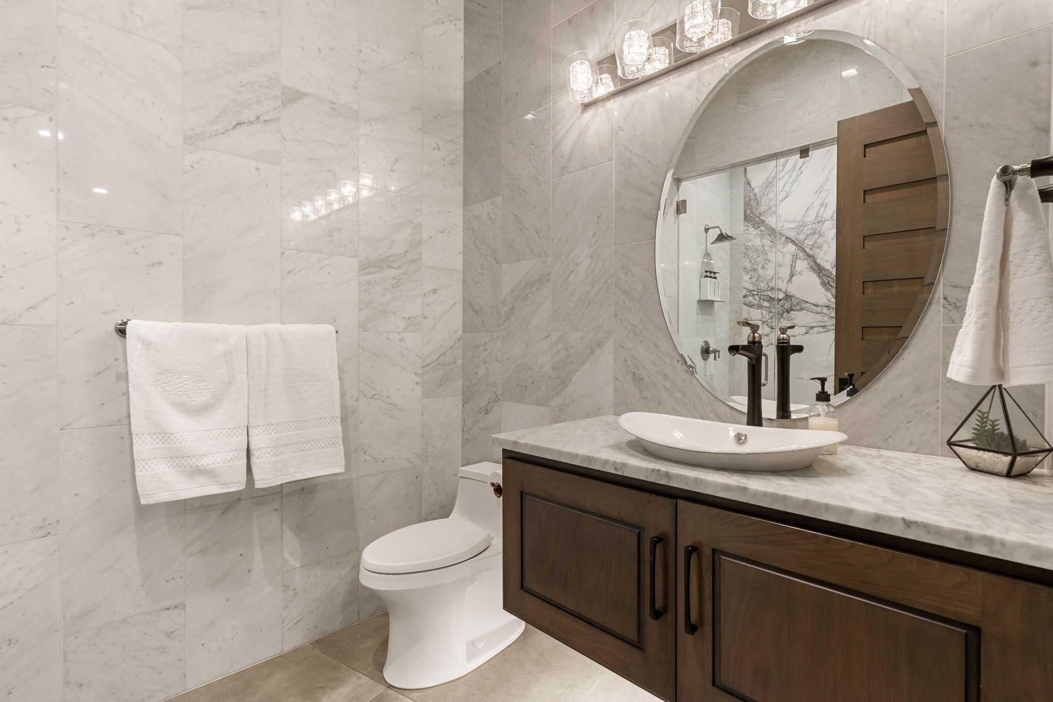 A modern bathroom with a marble floor, a white toilet, a wooden vanity with a sink, and a large mirror.
