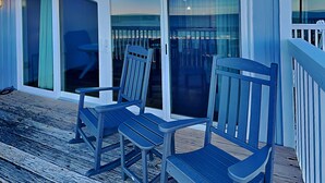 A wooden deck with two blue Adirondack chairs, a glass door leading to a covered area, and a white railing. - Sea Colony #35B - Sunrise Balcony +  Community Pool (Carolina Beach)