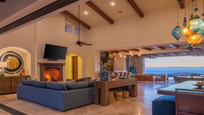 A spacious living area with a fireplace, a sofa, a dining table, and a view of the outdoors. - Modern Diamante Golf Villa w/ Heated Pool, Hot Tub, Golf Cart & Ocean Views (Cabo San Lucas)