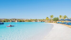 A clear blue pool with a kayaker, a sandy beach, and lounge chairs with umbrellas. - Modern Diamante Golf Villa w/ Heated Pool, Hot Tub, Golf Cart & Ocean Views (Cabo San Lucas)