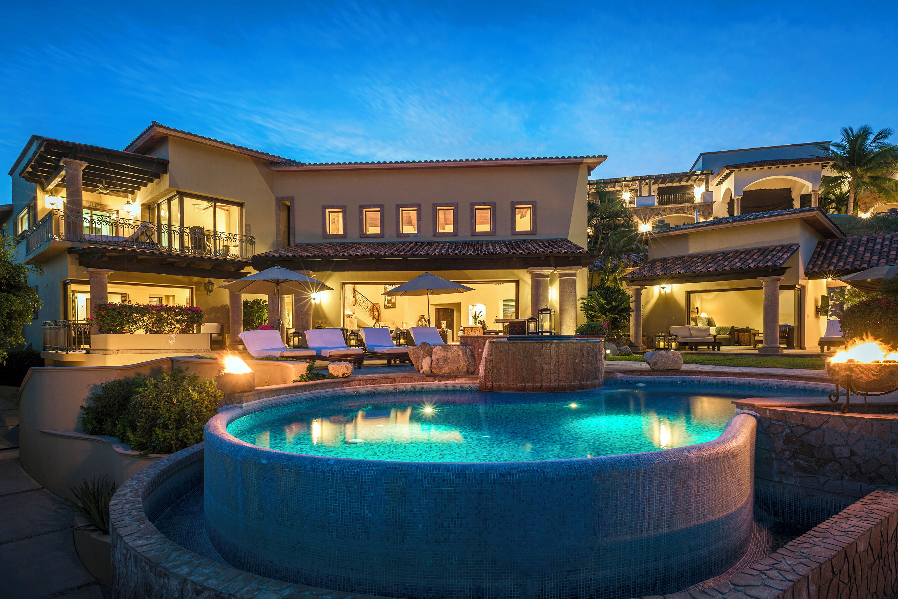 A luxurious pool area with a hot tub, lounge chairs, and umbrellas, surrounded by a well-lit, multi-story building with a Mediterranean architectural style.