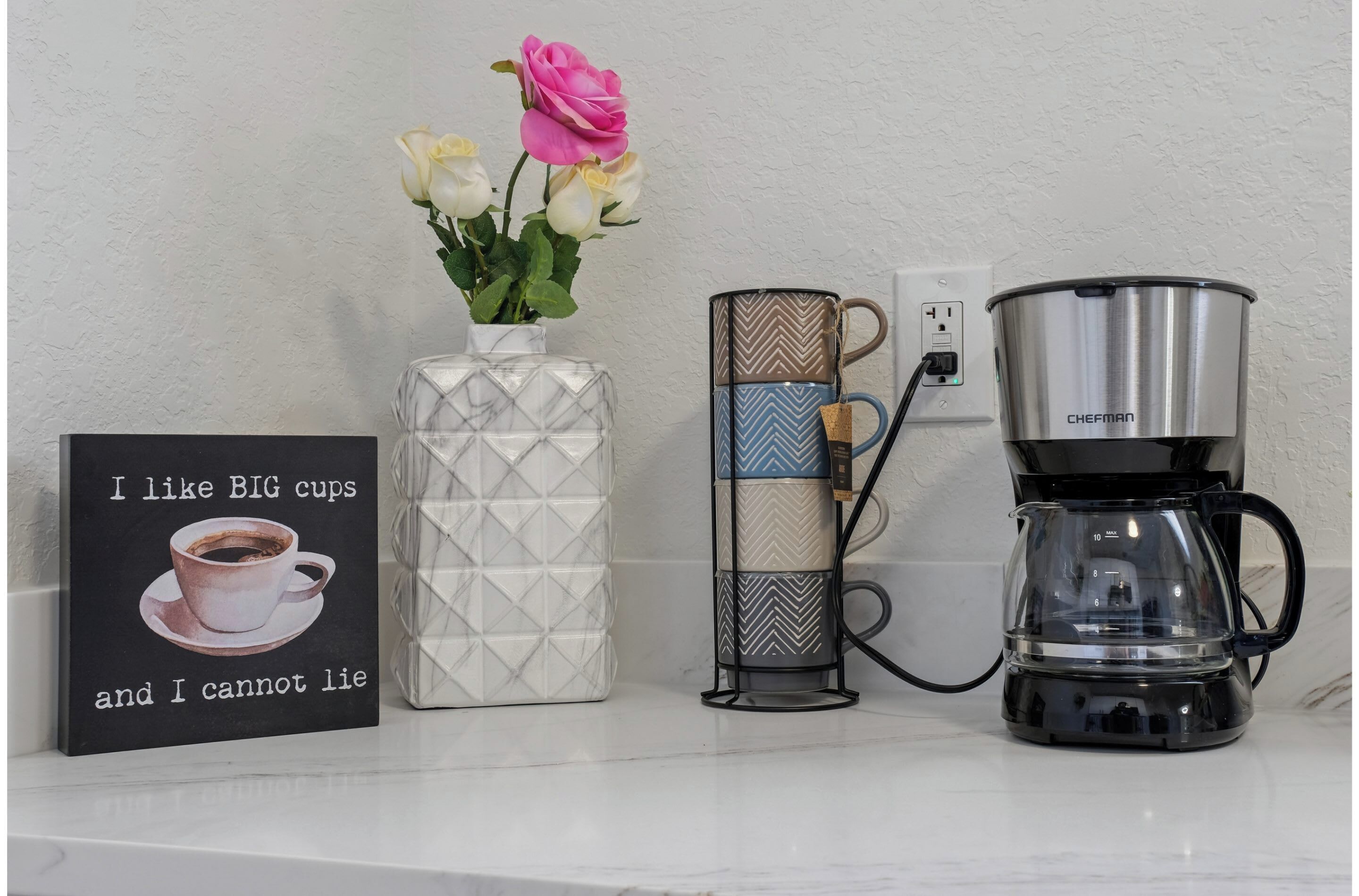 Un comptoir de cuisine avec une machine à café, une tasse, un vase de fleurs et un panneau.