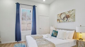 A neatly made bed with white bedding, a blue and white patterned rug, a white door, and a wall-mounted abstract painting. - Cozy Historic Gem: 5 Mins to Downtown SA (San Antonio)