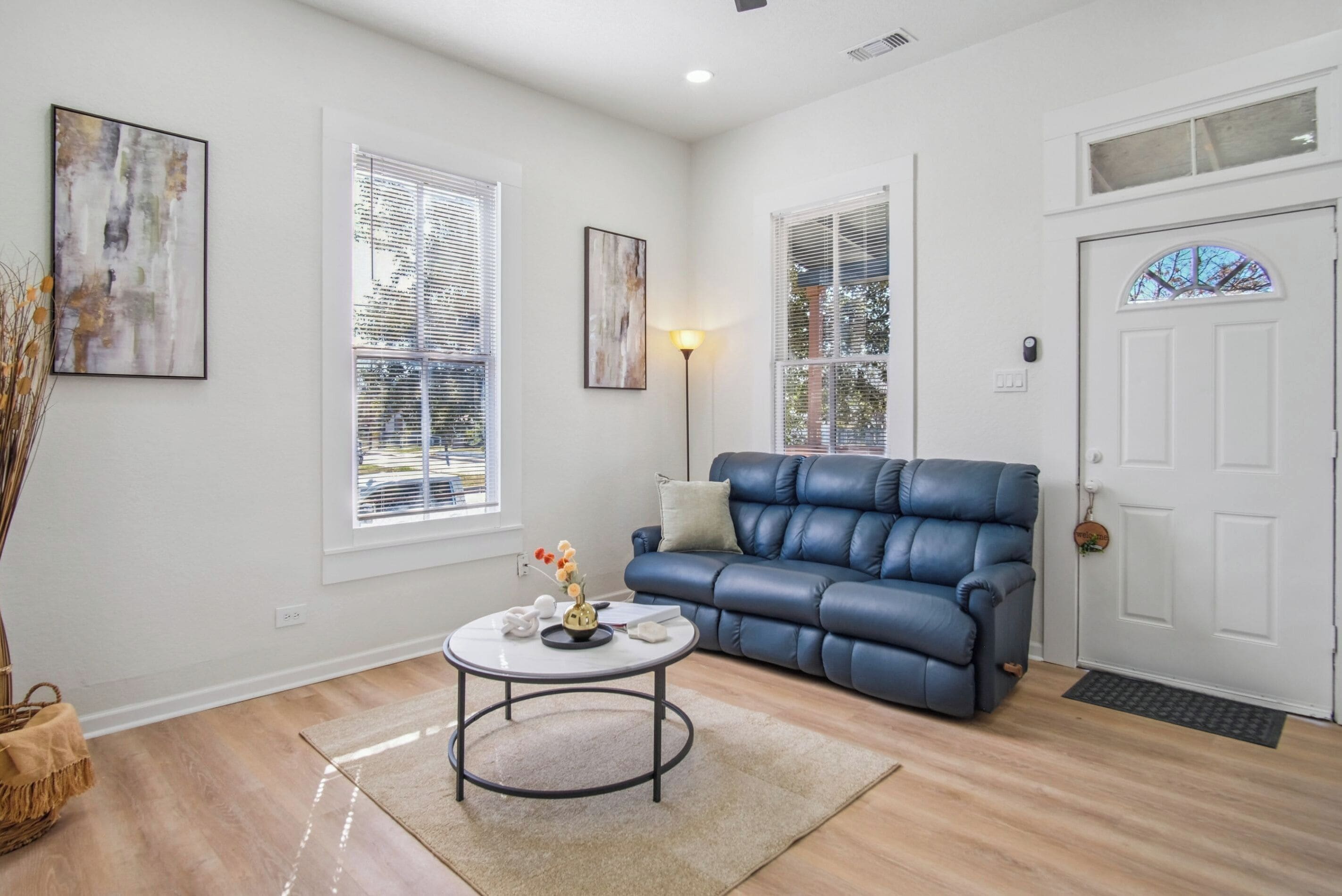 A living room with a blue leather sofa, a round coffee table, and two windows with blinds.
