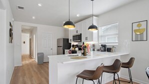 A modern kitchen with white cabinetry, a central island with stools, pendant lighting, and a view into the adjacent living area. - Minutes to Downtown: Spacious 4br/3ba Historic Gem (San Antonio)