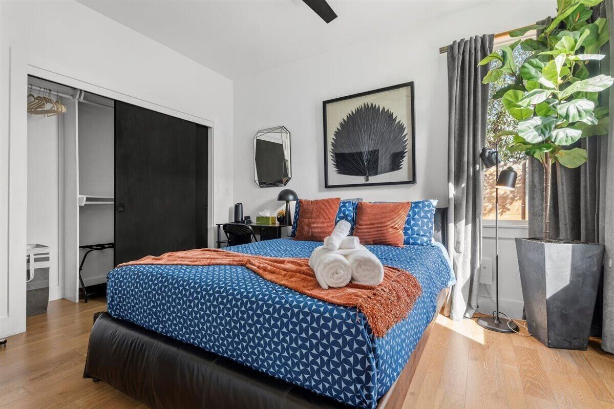 A bedroom with a bed, a mirror, a framed leaf artwork, a potted plant, and a door to a bathroom.