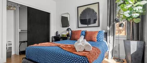 A bedroom with a bed, a mirror, a framed leaf artwork, a potted plant, and a door to a bathroom.