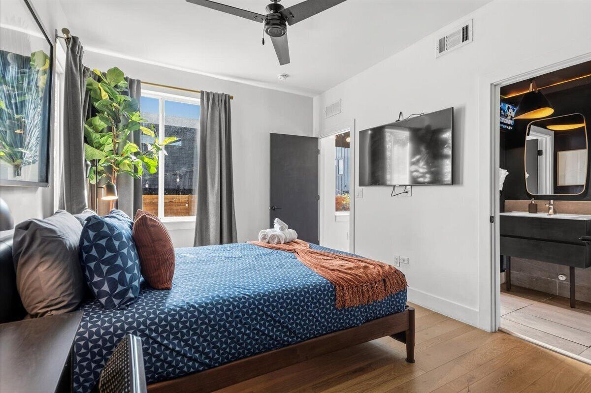 A modern bedroom with a bed, a ceiling fan, a large window with curtains, a wall-mounted TV, and a bathroom with a mirror and sink.