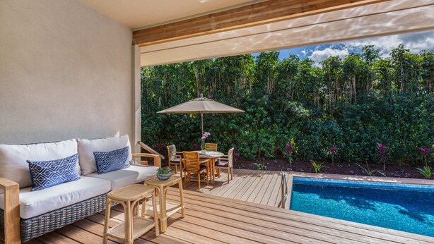 Una terraza de madera con piscina, un sofá con cojines estampados, una mesa, sillas y una sombrilla.
