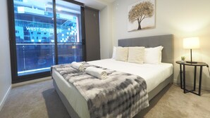 Ein Hotelzimmer mit großem Fenster, einem Bett mit grauem Kopfteil, einem Nachttisch mit Lampe und einem Baumgemälde an der Wand.