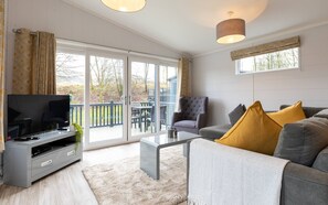 A modern living room with a grey sofa, a coffee table, and a TV on a grey stand. There is a balcony with a railing and a view of trees. - Landal Aysgarth (Leyburn)