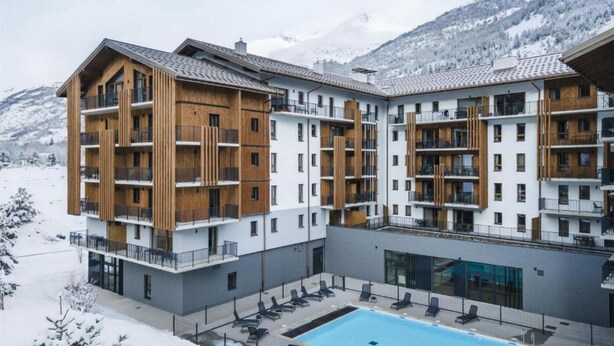 Een modern hotel met meerdere verdiepingen, houten details, een zwembad en een besneeuwde berg op de achtergrond.