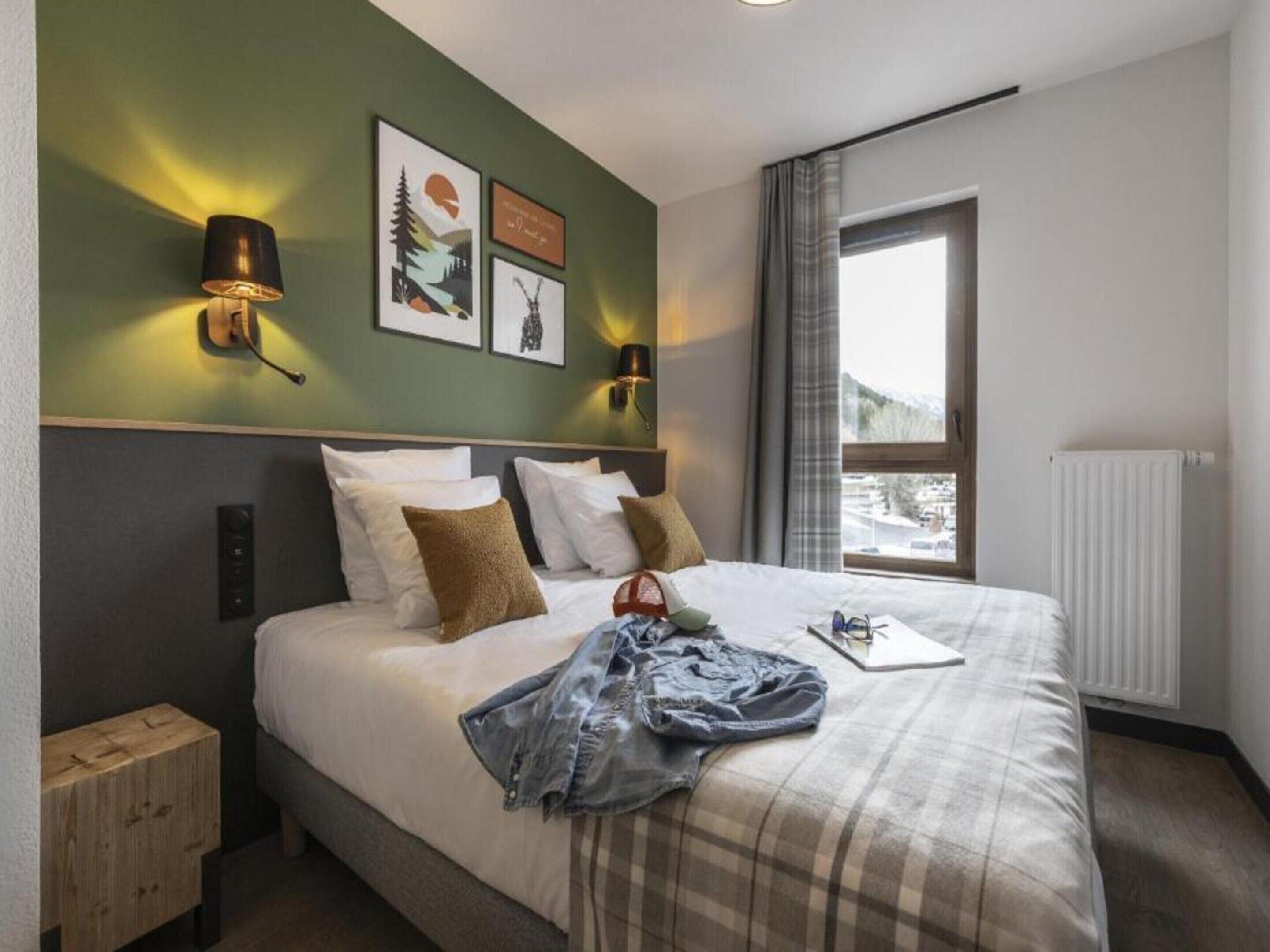 Hotellihuone, jossa on suuri sänky, kaksi seinään kiinnitettyä lamppua, ikkuna, josta on näkymä, ja patteri.