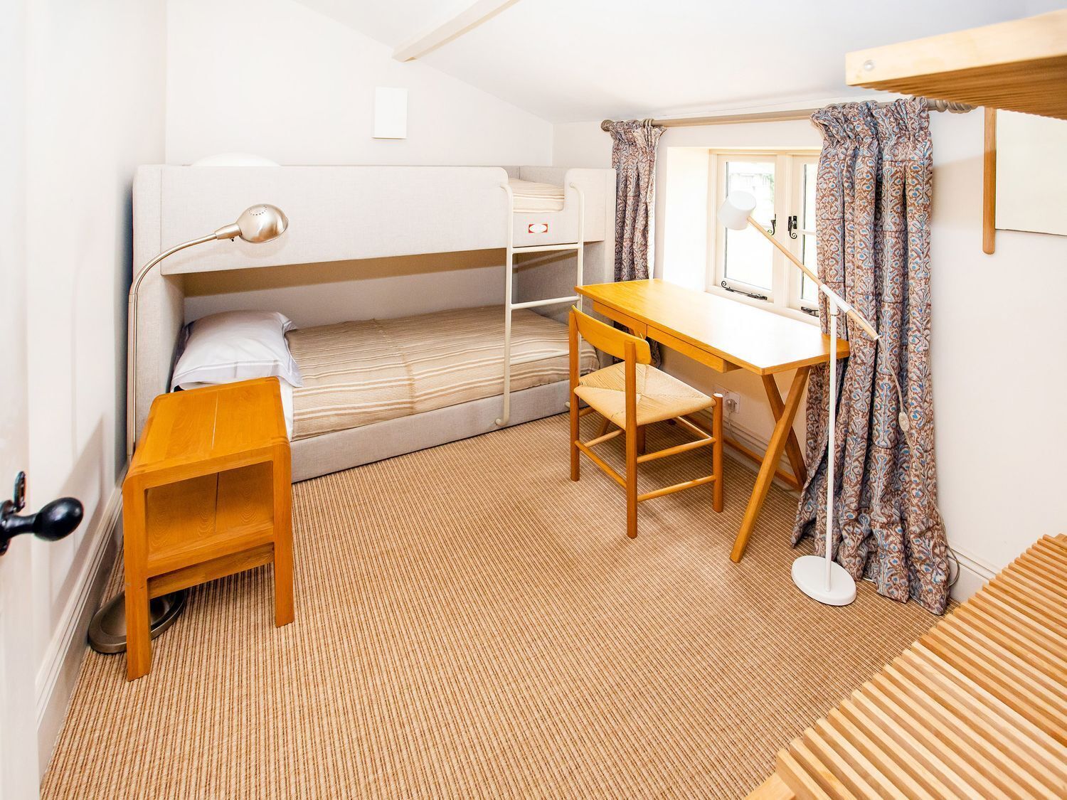 A compact dormitory room with bunk beds, a wooden desk, a chair, a small table, a lamp, and a window with patterned curtains.