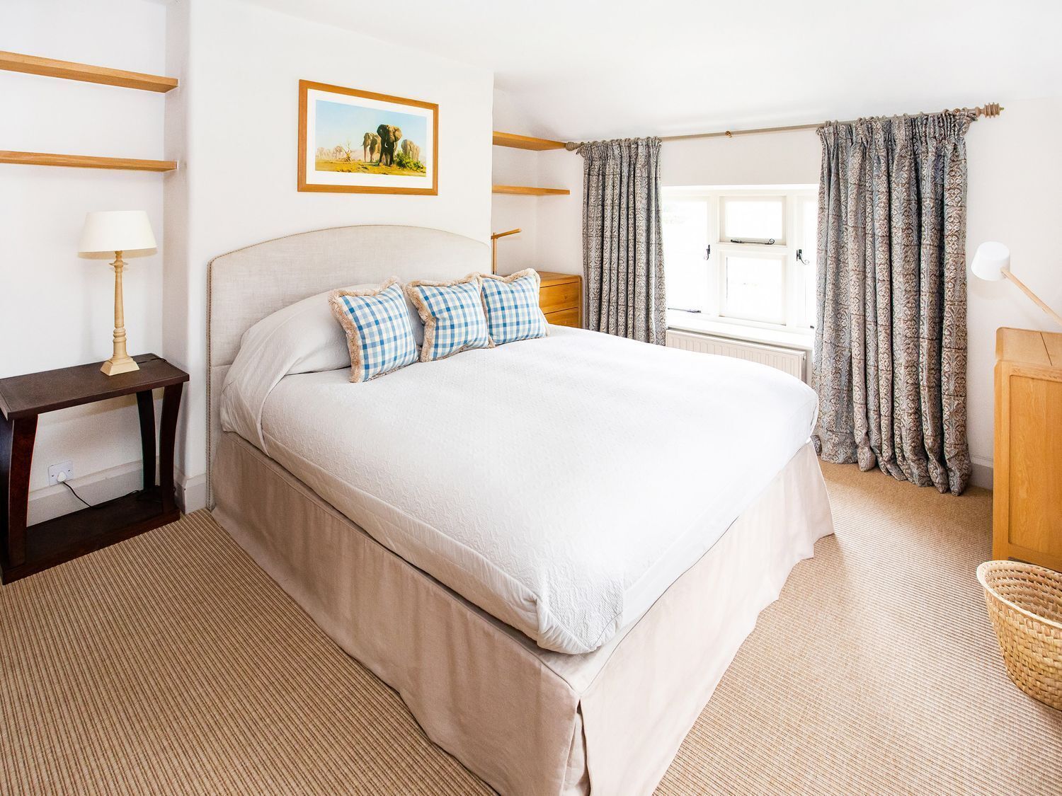 A neatly arranged bedroom with a large bed, a bedside table, a lamp, a framed picture on the wall, and patterned curtains.