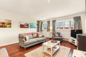 Ein modernes Wohnzimmer mit einer grauen Couch, einem weißen Couchtisch und einem Fernseher. An der Wand hängen drei Gemälde und ein Fenster mit Vorhängen.
