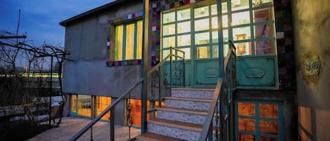 Un edificio con una escalera que lleva a una puerta con marco verde, una ventana con cortinas amarillas y una barandilla de metal.