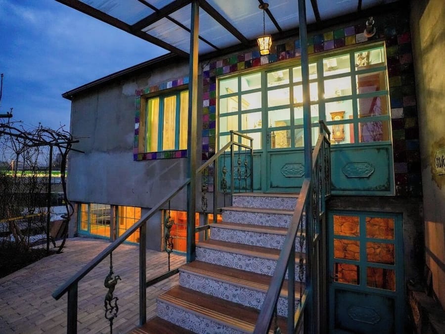A building with a staircase leading to a door with a green frame, a window with yellow curtains, and a metal railing.