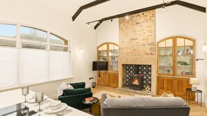 Ein gemütliches Wohnzimmer mit Kamin, einem grünen Sofa, einem Essplatz und einem großen Fenster mit Vorhängen.