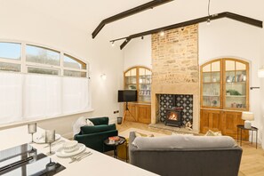 A cozy living room with a fireplace, a green sofa, a dining table set, and a large window with curtains. - The Reading Room - North East Escapes (Shilbottle)