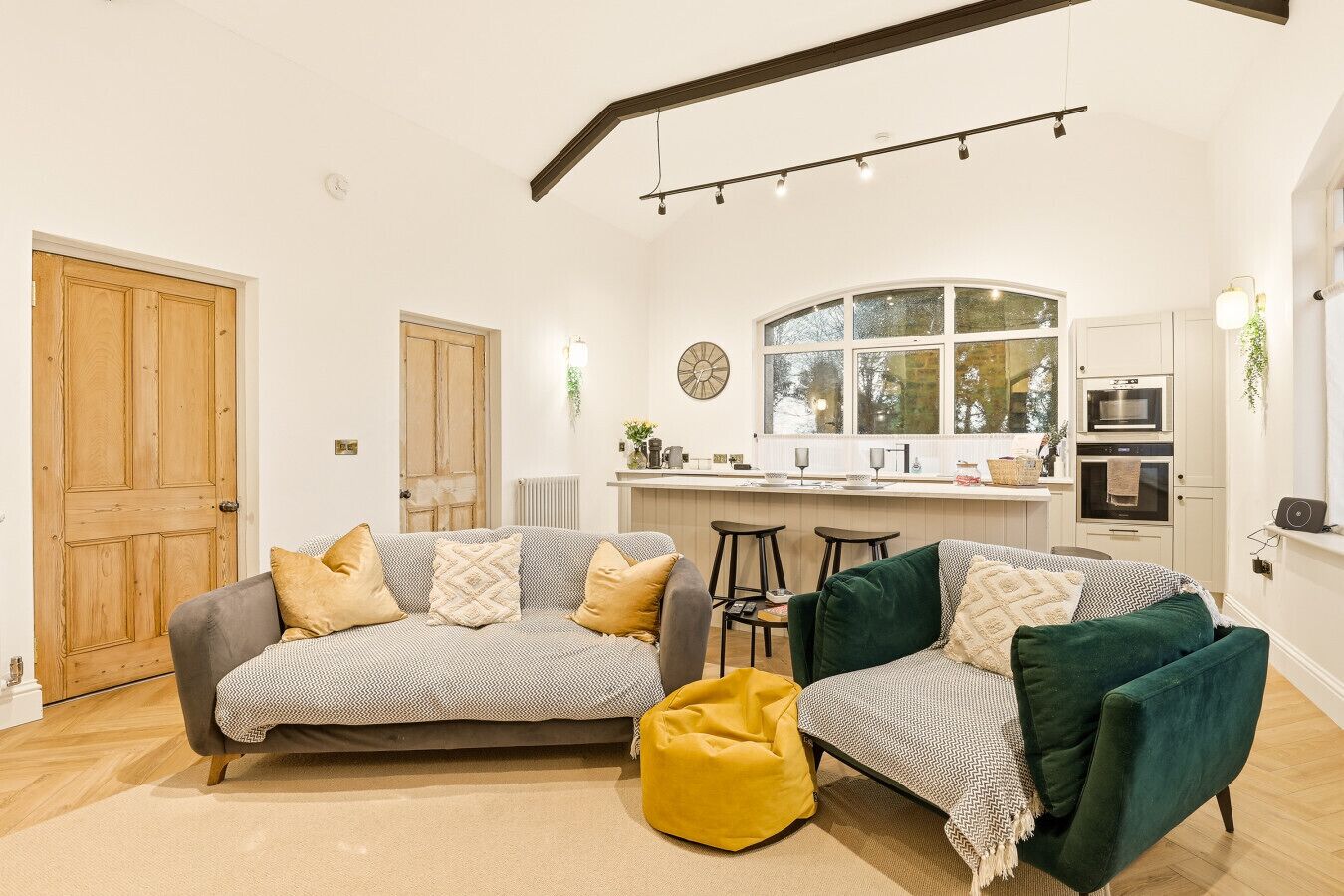 Una sala de estar moderna con un sofá gris, un sillón de saco amarillo y un sillón verde. Se ve un área de cocina con encimera blanca y taburetes de bar al fondo.