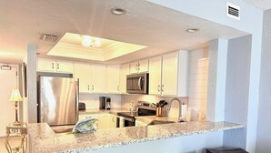 A modern kitchen with white cabinets, stainless steel appliances, a granite countertop, and bar stools. - Feel the Sea Breeze, Stylish 2 BR Condo on the Beach W/ Pool (Daytona Beach)
