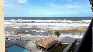 A pool area with lounge chairs and a thatched-roof structure, overlooking a beach with waves and a clear sky. - Feel the Sea Breeze, Stylish 2 BR Condo on the Beach W/ Pool (Daytona Beach)