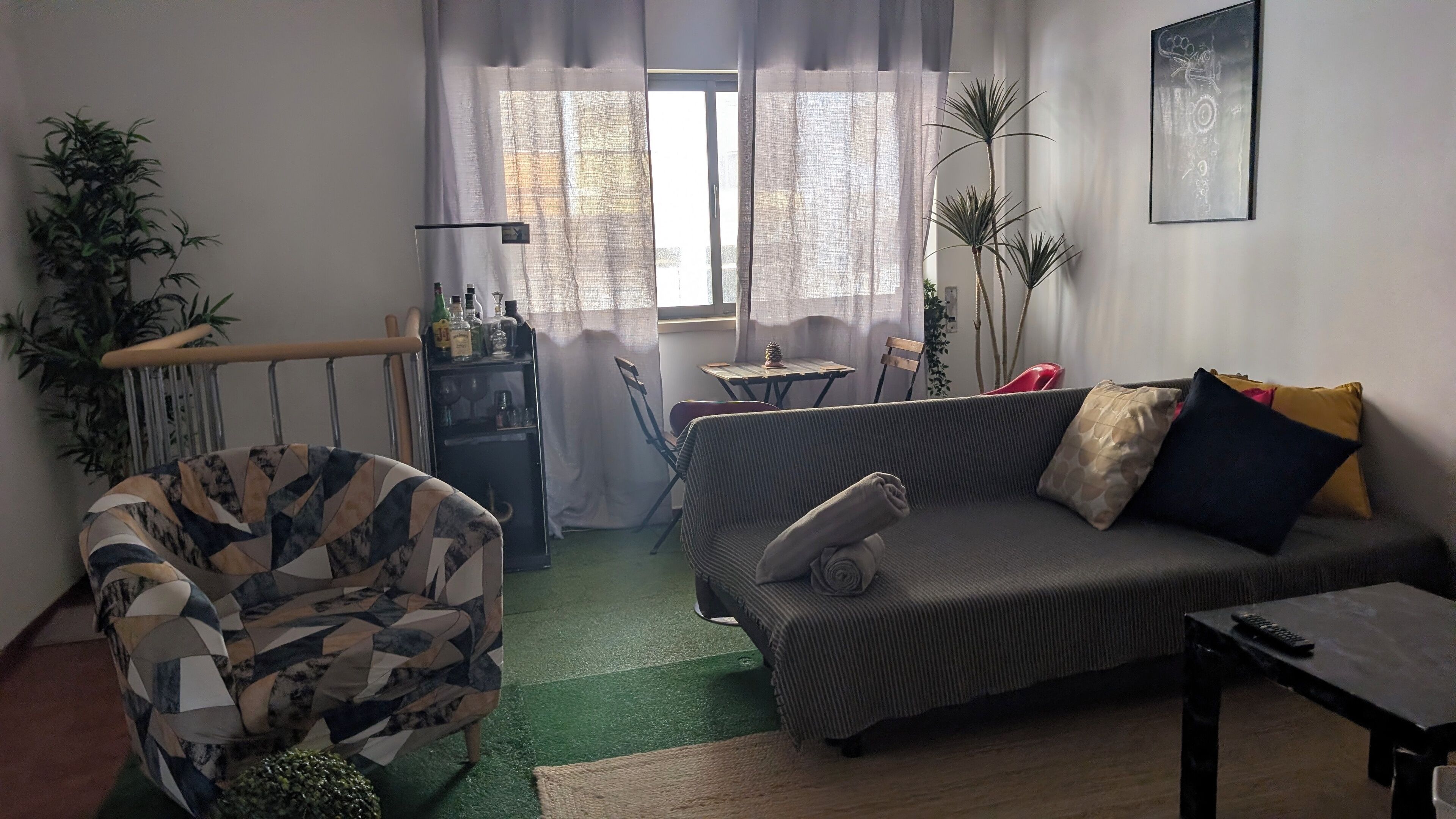 Un salon avec un canapé, un fauteuil et une petite table. Il y a une fenêtre avec des rideaux, une plante et une étagère avec des bouteilles.