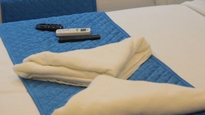 A bed with a blue quilted cover, a folded white towel, and two remote controls on the bed. - Niharika Heights Luxury Hotel (Bengaluru)