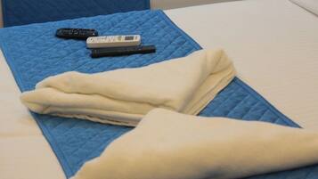 A bed with a blue quilted cover, a folded white towel, and two remote controls on the bed.