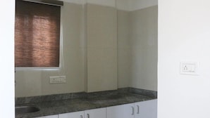 A kitchen with white cabinets, a granite countertop, a window with blinds, and a wall socket. - Niharika Heights Luxury Hotel (Bengaluru)