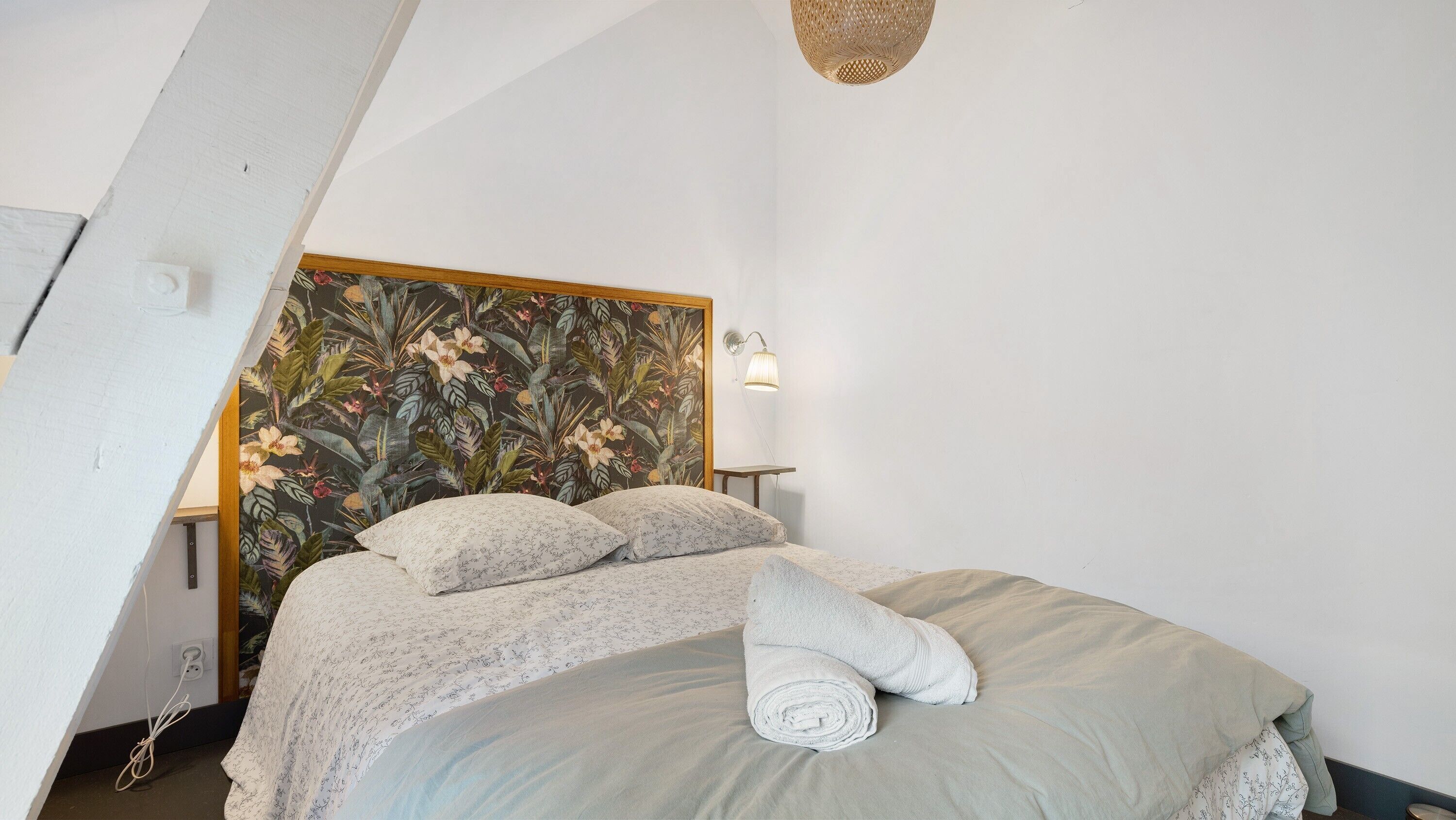 Ein Schlafzimmer mit einem Bett, einer Blütenmuster-verzierten Kopfteil, einer Wandleuchte und einem aufgerollten Handtuch auf dem Bett.
