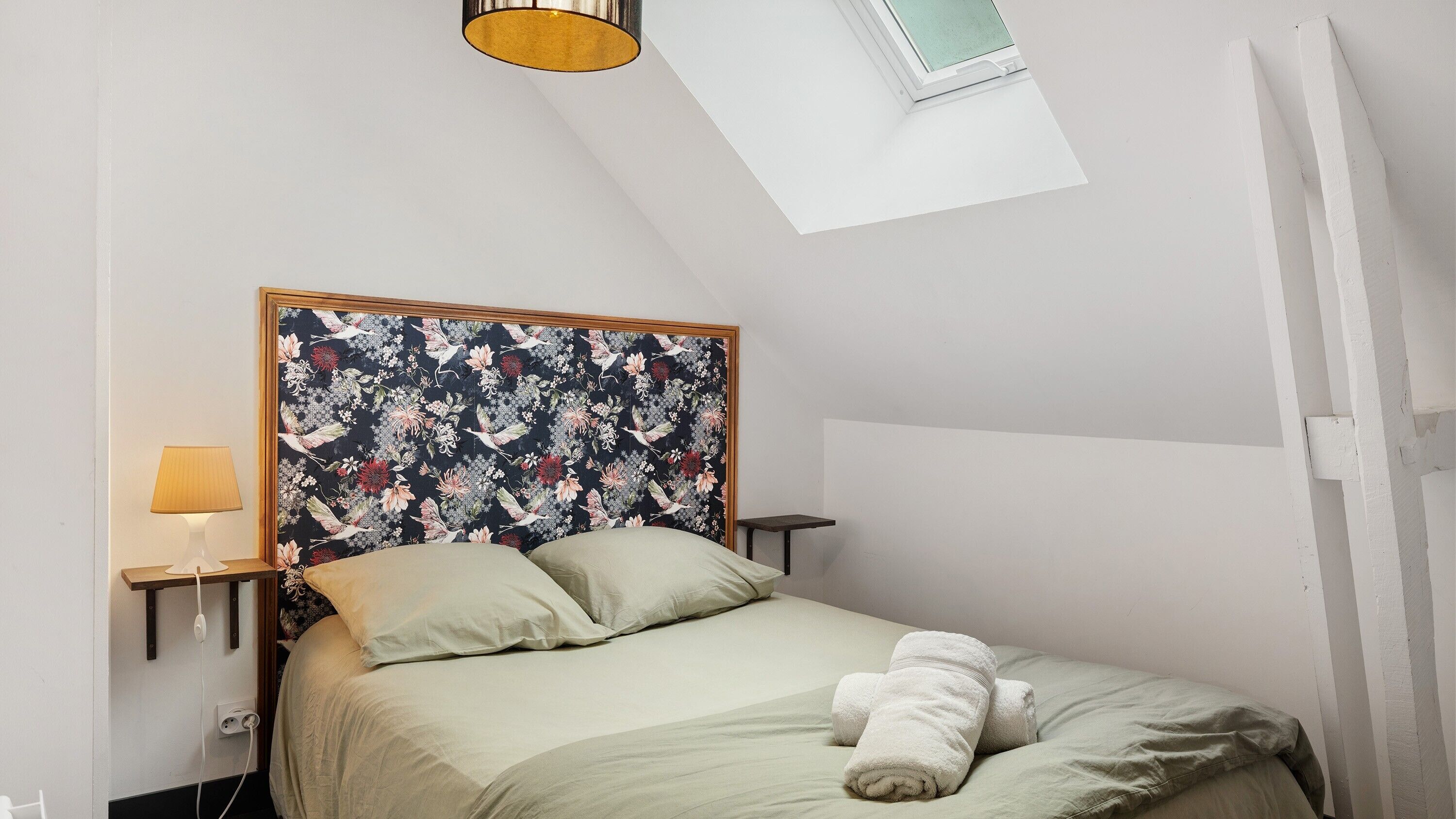 A bedroom with a bed, a nightstand, a lamp, a window, and a skylight.