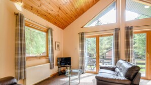 A cozy living room with a wooden ceiling, large windows, a leather sofa, a glass coffee table, and a television.