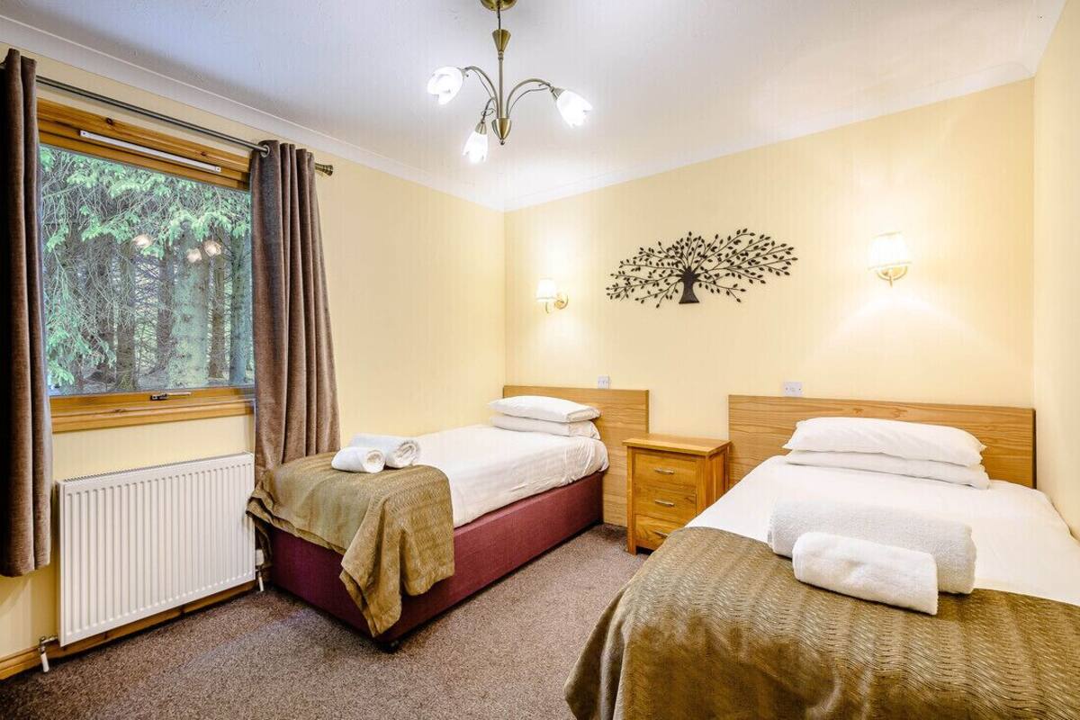 A hotel room with two beds, a window with curtains, a radiator, a wooden nightstand, and a wall-mounted light fixture.