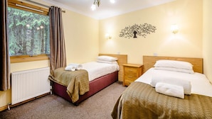 A hotel room with two beds, a window with curtains, a radiator, a wooden nightstand, and a wall-mounted light fixture.