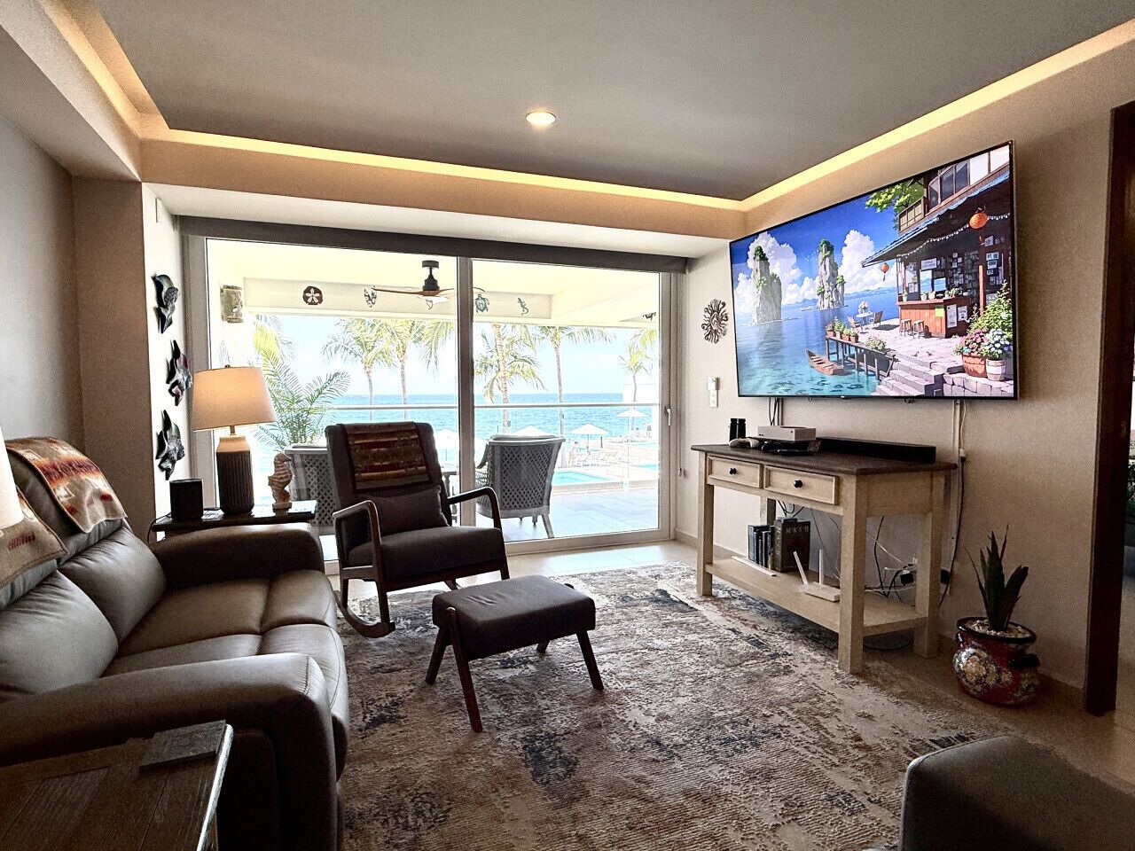 Un salon comprenant un canapé, un fauteuil, un pouf, une télévision et offrant une vue sur l’océan grâce à de grandes fenêtres.